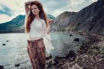 Beautiful boho styled model wearing white crochet swimsuit posing on the beach in sunlight. Red-haired girl with freckles. Crimea, Fox bay, Koktebel