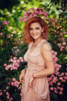 Lovely young woman in a pink dress and red hair, waiting for a date. A walk through the summer city, pink flowering bushes