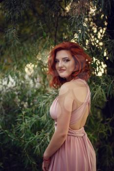 Lovely young woman in a pink dress and red hair, waiting for a date. A walk through the summer city, pink flowering bushes