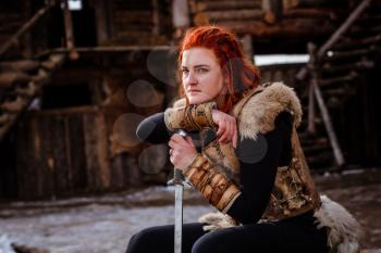 Red-haired woman is a Viking. Against the backdrop of a village or an ancient settlement. In the hands of a sword