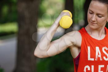 Sport training concept. The girl is engaged in fitness in nature. Side view of young woman doing stretching exercise using dumbbells. Active athlete working out on fitness routine to lose weight.