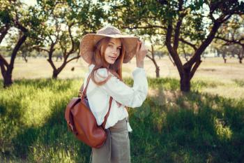 young girl in a straw hat travels green meadows. Beautiful red-haired hipster woman in a green garden