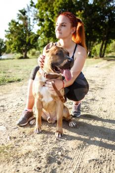 redhair jouful young woman caressing their dog, wearing sport clothing, enjoying their time and vacation in sunny park