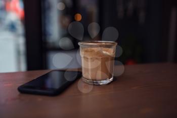 Close up of mobile phone lying on a wooden table near cup of cappuccino in coffee shop interior, leisure time concept, rest in cafe