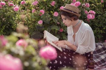 beautiful girl wearing hat with book sitting on grass in rose gaden. A country girl enjoys the sunset, wine and reading books in the fragrant rose garden. The concept of perfume