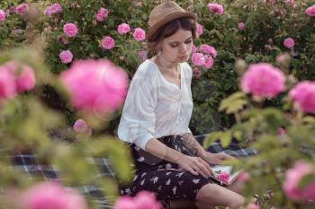 beautiful girl wearing hat with book sitting on grass in rose gaden. A country girl enjoys the sunset, wine and reading books in the fragrant rose garden. The concept of perfume