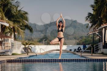 young woman practicing pose yoga on swimming pool and the nature beach at sunset or sunrise. Yoga and Healthy Concept. yoga in tree pose
