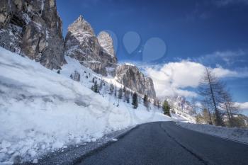 Scenic winter view from the asphalt road in the mountains covered with snow and trees on the side of the road on a background of blue sky and clouds. South Tyrol, North of Italy, Gardena Pass