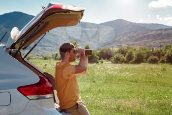 Young man at mountain looking through binoculars sitting on the trunk of a car. Banner with copy space. idea and concept of travel and discoveries, adventures and freedom
