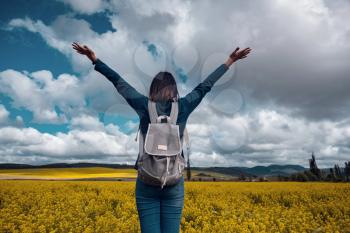 Woman traveler in a blue sweater and jeans in a flowering rapeseed field with hands up and looking towards the blue sky. Happy beautiful young woman enjoys blooming rapeseed field