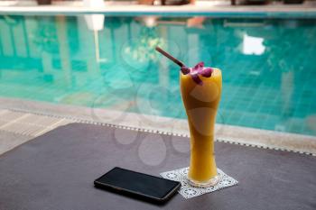 Smartphone and mango smoothie in a tall glass on a sunbed near the swimming pool. Idea and concept of vacation, travel and discoveries