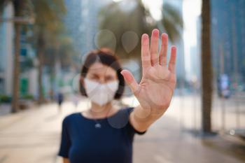 Portrait of young woman wearing in blue dress and white mask for prevent virus, walk in front of skycrapers in modern city. Gesture stop , focus on the hand