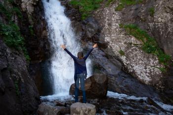 Young happy woman enjoing the waterfall. Woman standing in front of waterfall with rased hands and smile. Concept of ecotourism travel. Waterfall Shumka, Dombai, Karachay-Cherkessia, Russia