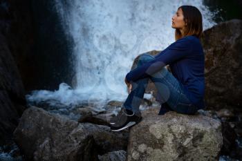 Beautiful woman hiker sitting near waterfall. Ecotourism concept image with happy woman. Mixed race Asian Caucasian traveler. Waterfall Shumka, Dombai, Karachay-Cherkessia, Russia
