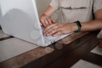 Focused young man wearing glasses using laptop, typing on keyboard, writing email or message, chatting, shopping, successful freelancer working online on computer, sitting in kitchen, closeup on hand