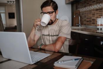 Young entrepreneur sitting at table drink coffee work at laptop thinking of problem solution, thoughtful male employee pondering considering idea looking at computer screen making decision