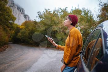 Woman beside a car and looking at smart phone surrounded autumn forest in mountains. Portrait of romantic hipster female, Warm autumn weather, calm scene. Wanderlust photo series.