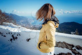 Beautiful Young Smiling Girl in her Winter Warm Clothing. on the background of a beautiful winter mountain landscape in nature, on a beautiful observation point in the mountains with sea views