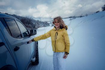 Young cheerful woman posing against the car and snow forest. Attractive woman dressed yellow jacket sitting at Passenger seat in silver car, winter season