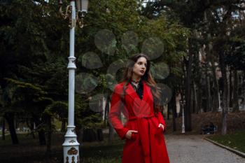 Beautiful young woman in red coat is enjoying autumn park. A pleasant walk on the background of park