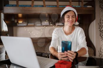cute young woman sit couch hold telephone look camera smiling wear white pullover in decorated living room indoors. Merry Christmas Covid 19 coronavirus social distance concept.