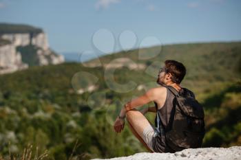 Young male hiker with backpack relaxing on top of a mountain during calm summer sunset - scenery from vacation. Tired traveling man resting on hill.