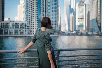 Happy young female traveler in the big city of Dubai, famous place Dubai marina. Luxury and comfortable tourism season in United Arab Emirates. Back or rear view of young woman in dress