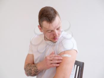 Man showing his vaccinated arm. Guy received a corona vaccine looking at plaster on his shoulder on white background.