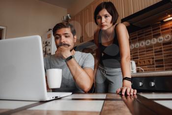 Young couple using laptop and smartphone in the kitchen, smiling man working online at home, family morning lifestyle with gadgets or devices addiction concept. readings news