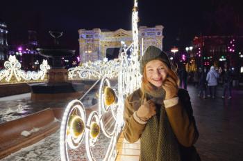 young smiling woman posing on the street. Festive Christmas fair in the background. Model wearing a stylish winter coat, knitted cap, scarf. Feeling happy in big city. Spending winter vacations Moscow