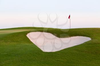 Heart shaped sand bunker in front of red flag of golf hole on beautiful course at sunset illustrating love for game of golf