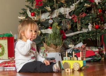 Young caucasian toddler sits in front of a Christmas tree opening gifts and toys  with a thoughful expression
