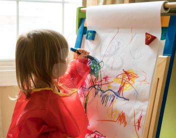 Young two year old girl drawing with crayon on paper mounted on an easel during playtime