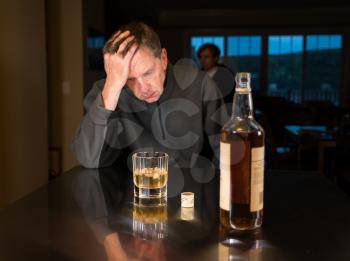 Senior adult male facing a kitchen table with alcoholic drink and looking very sad and depressed as wife is seen in the background