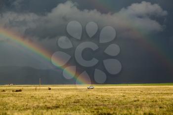Sun illuminating prairie grassland with dark and forbidding thunderclouds and downpour with van riding along lonely country road and rainbow ending over the vehicle