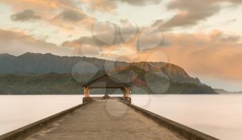 Rising sun illuminates the peaks of Na Pali mountains over the calm bay and Hanalei Pier in long exposure photo