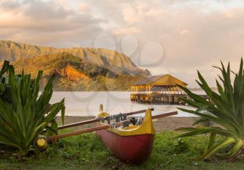 Red and yellow hawaiian canoe with outrigger on the beach at Hanalei pier at dawn as the sun lights the sky over Na Pali mountains