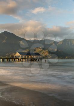 Swimmer stands in the ocean as the rising sun illuminates the peaks of Na Pali mountains over the calm bay and Hanalei Pier