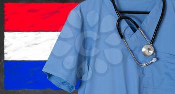 Blue doctor scrubs shirt and stethoscope hang empty in front of Luxembourg flag. Illustration of medical staff coming from other countries to staff health systems
