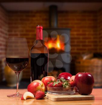 Red wine in bottle and glass with apple and strawberries on wooden table in front of roaring fire inside wood burning stove in brick fireplace