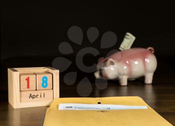 Wooden letters in calendar with Form 1040 income tax for 2016 showing tax day for filing is April 18 2017 with savings piggy bank in background