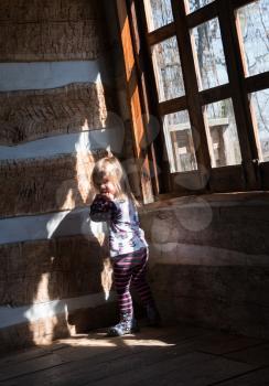 Small and lonely caucasian baby girl or toddler standing inside empty old wooden cabin to suggest poverty or recession