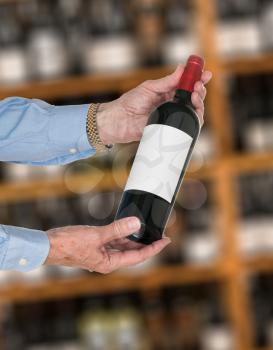 Senior caucasian wine steward offering a bottle of red wine with blank label to a customer in a wine store or winery. The bottle is ready with copy space.