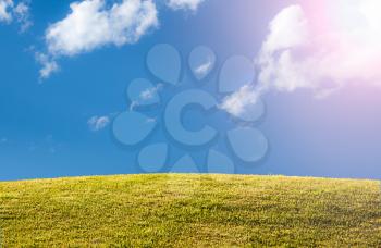 Bright green sunlit grass hill with blue sky and clouds with sun flare and copy space