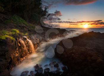 Setting sun at sunset illuminates a small waterfall falling into the ocean with the Na Pali coastline by Hanalei in the distance