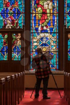 Photographer takes photo from the aisle between pews illuminated by light from stained glass window