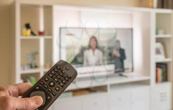 Senior male hand holding TV remote in bright modern apartment with large screen TV
