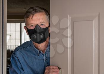Senior man with face mask looking worried at front door in case visitors bring coronavirus to his home