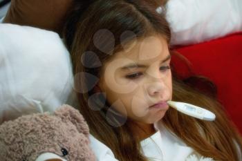A small ill girl lies in a bed covered with a blanket next to her favorite toy bear and holds in her mouth an electronic thermometer.