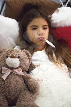 A small ill girl lies in a bed covered with a blanket next to her favorite toy bear and holds in her mouth an electronic thermometer.